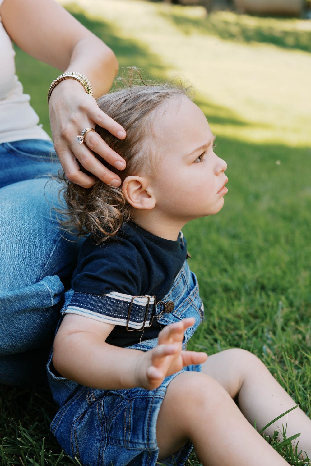 Tender moment of mother gently stroking curly-haired toddler sitting on green grass outdoors.