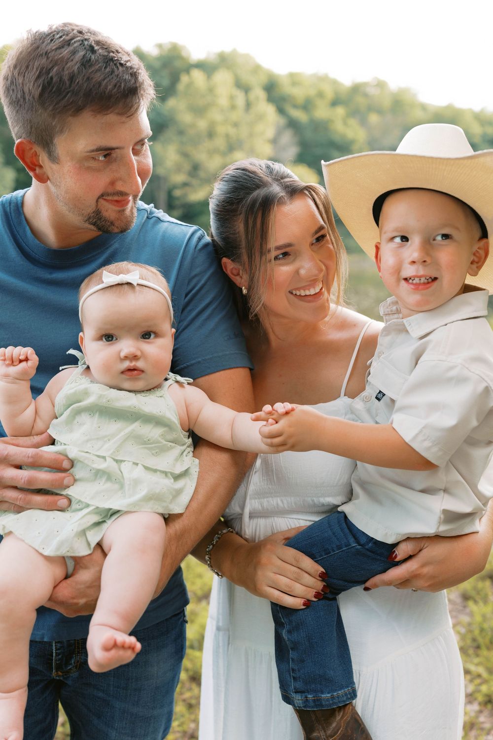 Happy family of four posing outdoors, parents holding baby girl and toddler boy in cowboy hat.