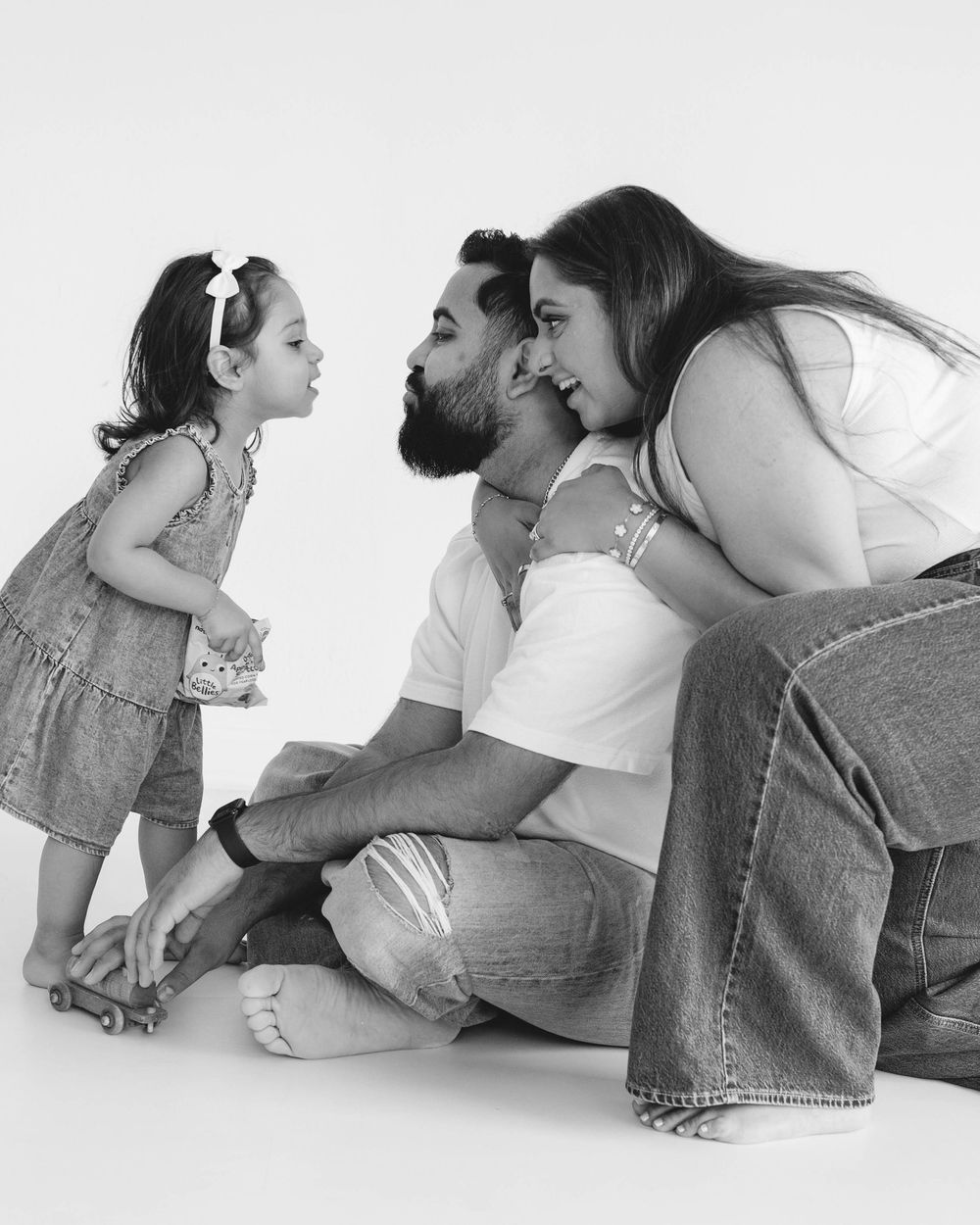 Black and white family photo of parents and toddler girl playing with toy on white studio background.
