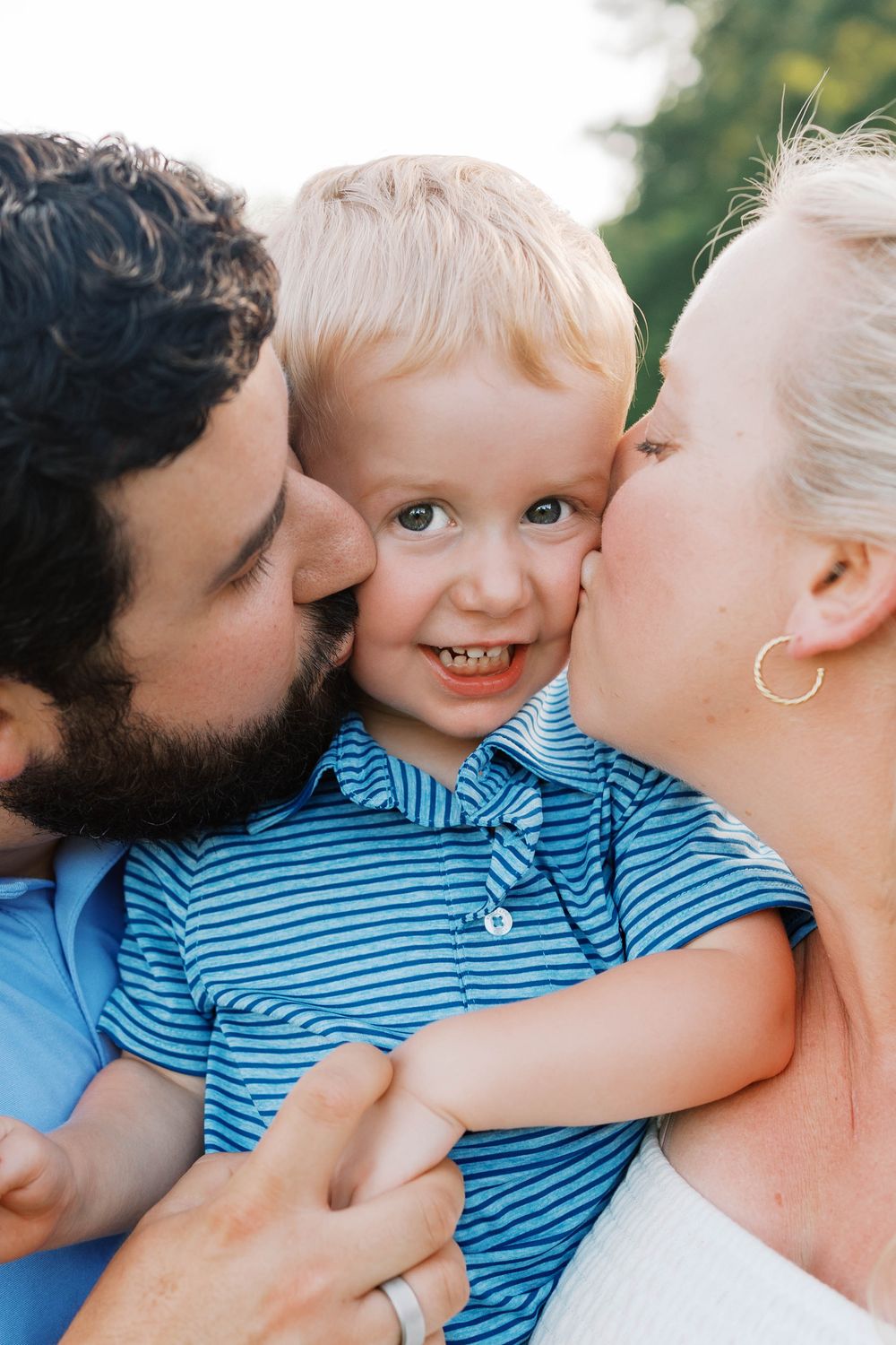 Loving parents simultaneously kissing smiling blonde toddler boy on both cheeks outdoors.