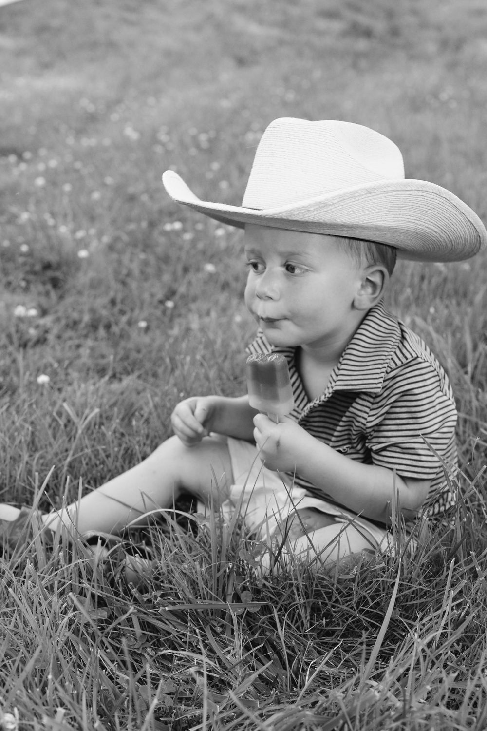 Cute young boy in cowboy hat sitting in grass eating popsicle, black and white photo.
