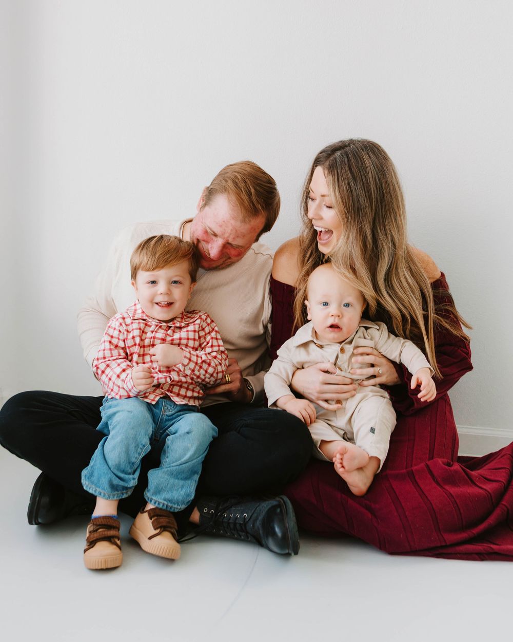 Joyful redhead family of four sitting on floor, parents cuddling two young boys in studio portrait.