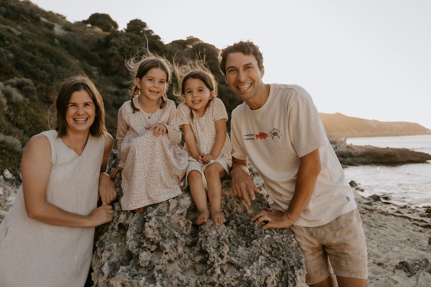 Family of four smiling on rocky beach at sunset, two girls sitting on rocks between parents in casual white outfits.