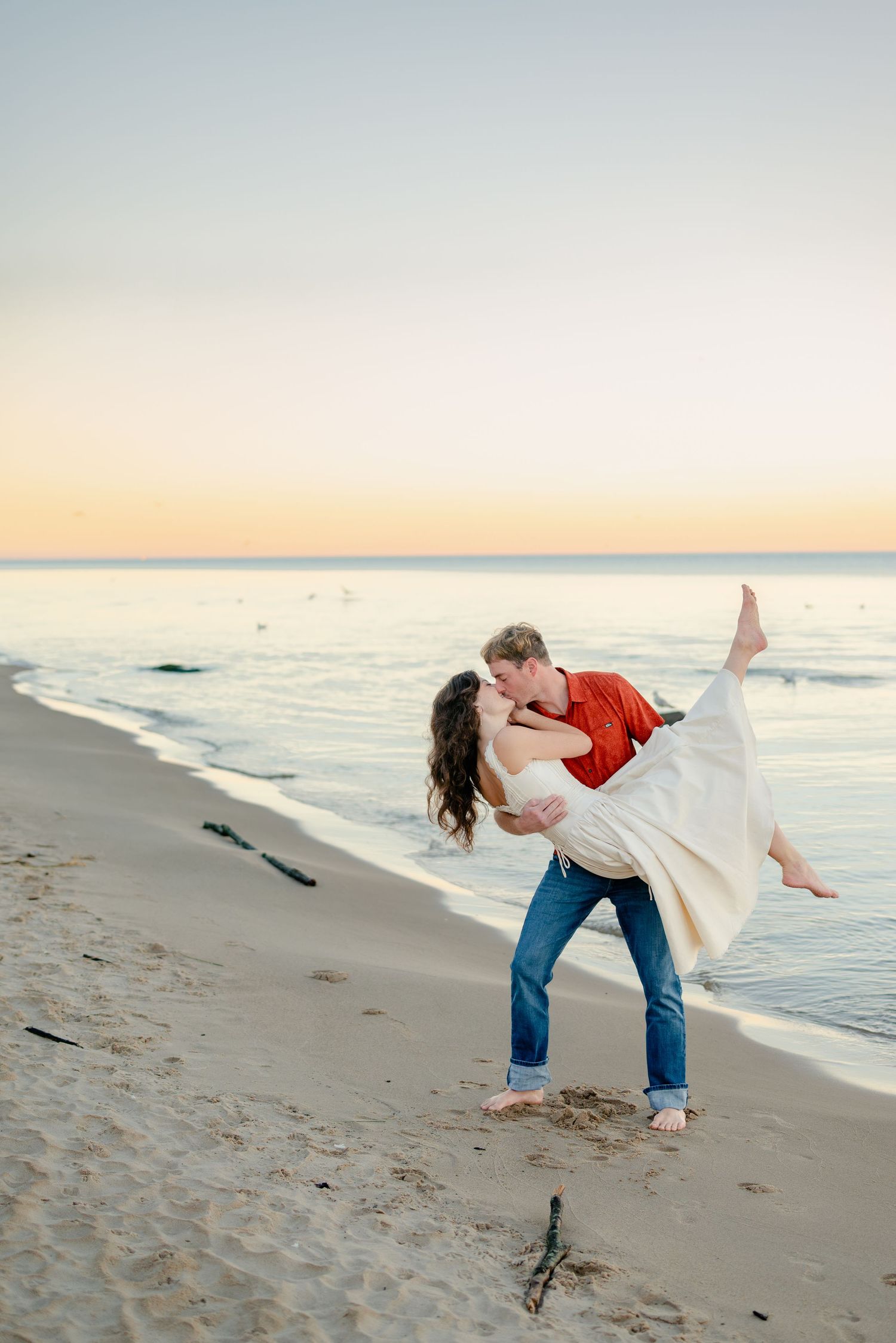 Couple sharing a romantic dip kiss on a sandy beach at sunset, man in red shirt and jeans lifting woman in white dress.