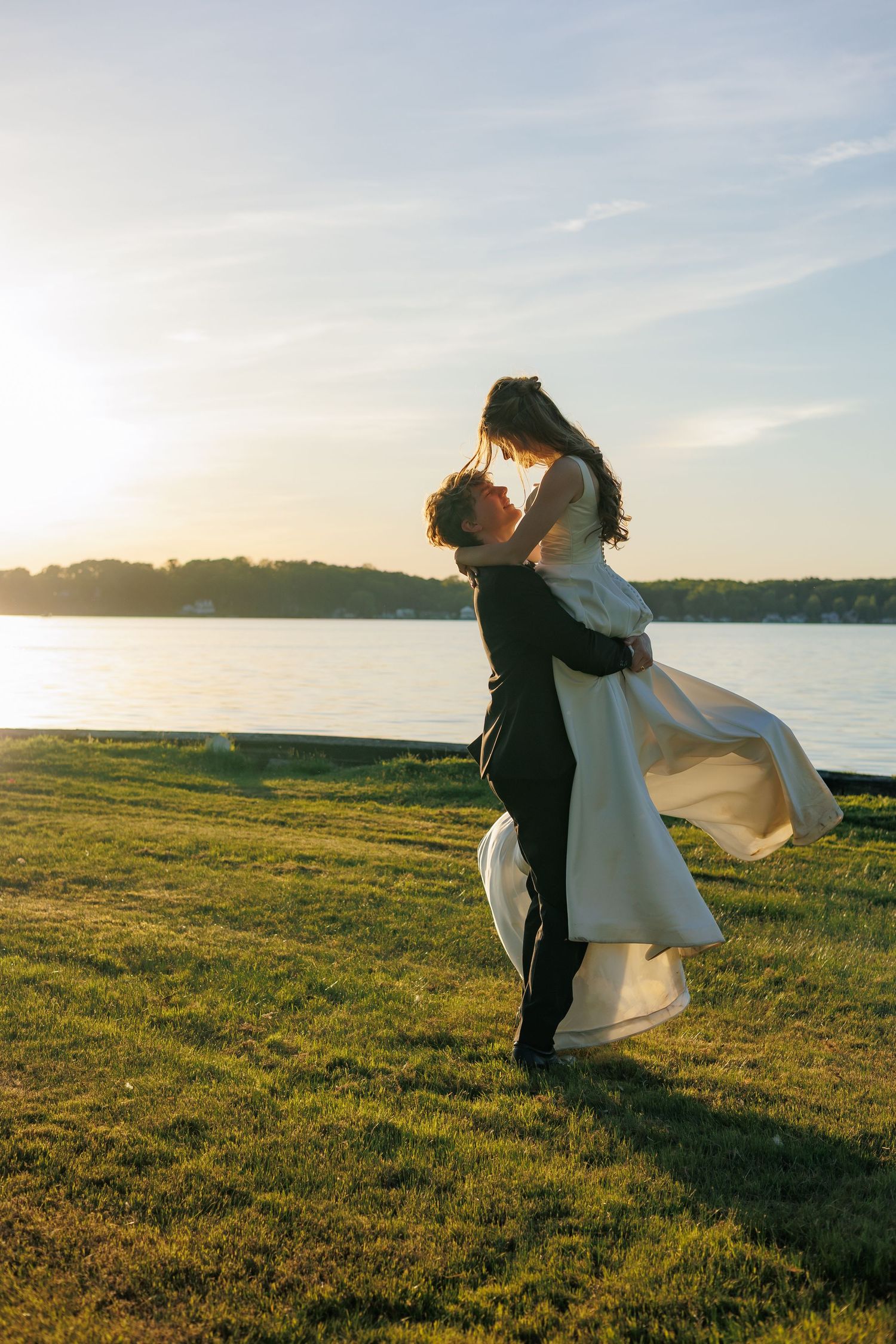 Black and white photo of a couple kissing in wedding attire by a sunlit lake near a willow tree.