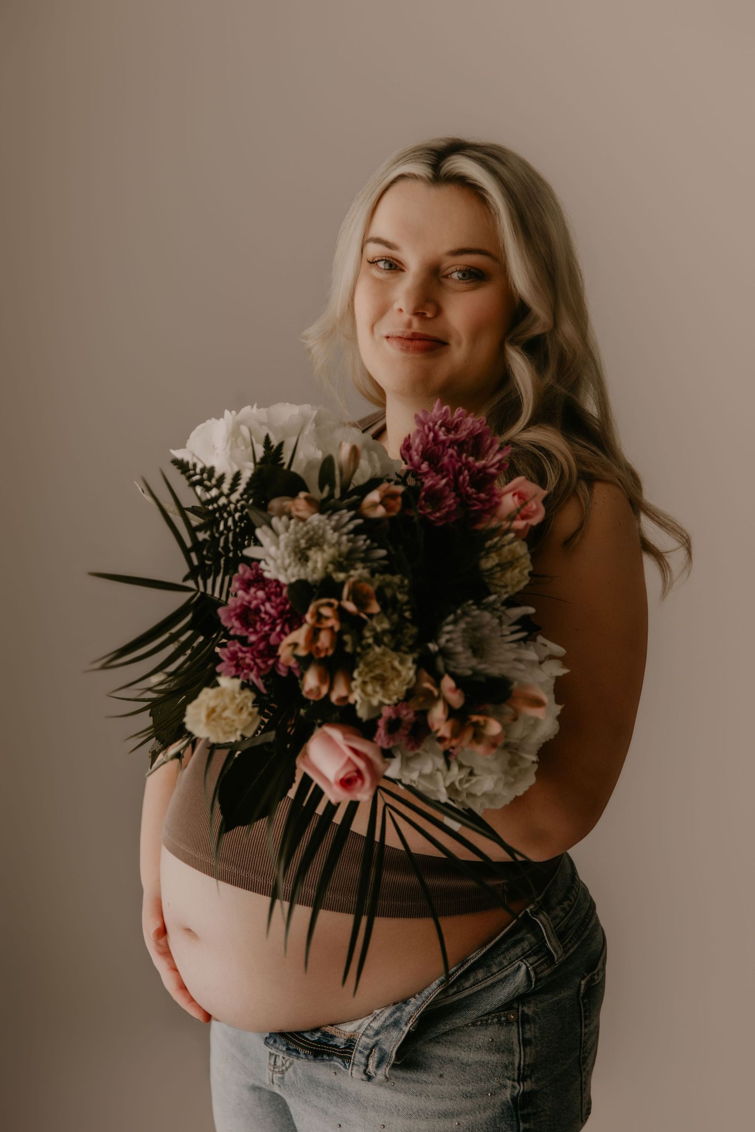 Pregnant woman holding a bouquet of pink and white flowers, wearing jeans and a sheer top, posing against a neutral background.