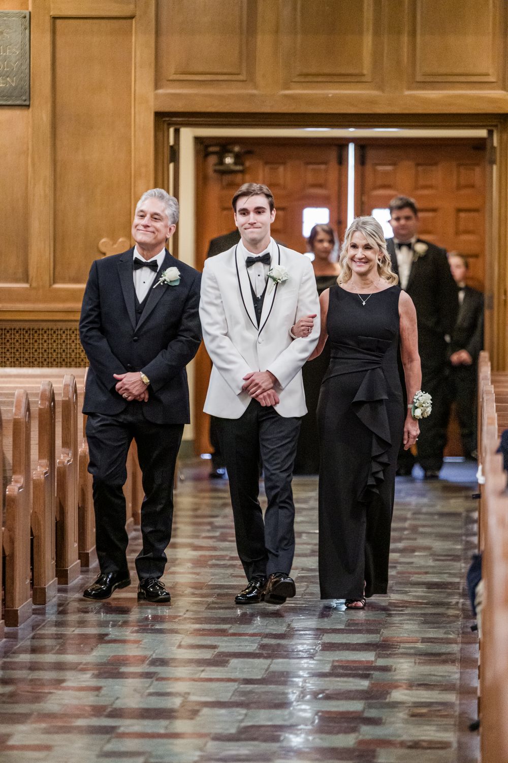 Groom in white tuxedo walks down church aisle with parents in black formal attire during wedding ceremony.