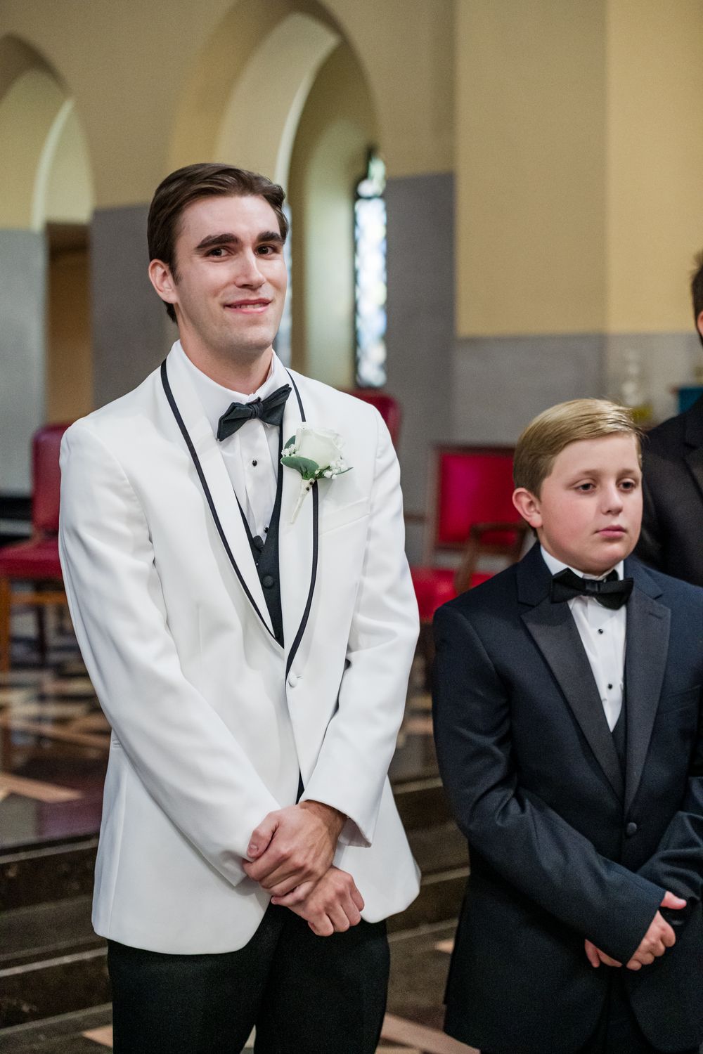 Emotional groom in white tuxedo jacket with boutonniere stands at church altar beside young boy in black tuxedo.
