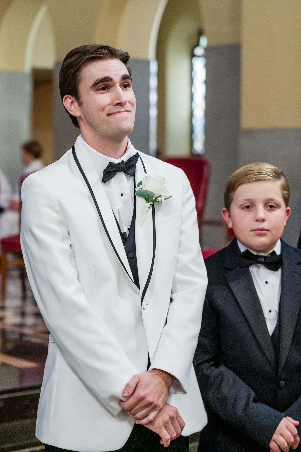 Groom in white tuxedo jacket with boutonniere smiling emotionally at altar beside young boy in black tuxedo in church.