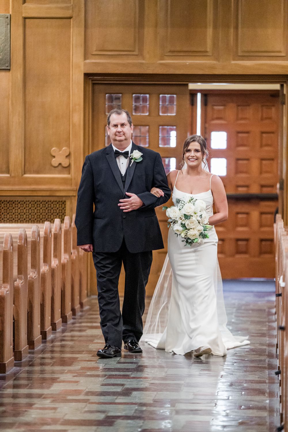 Bride in white gown walks down church aisle with escort in black tuxedo, holding white floral bouquet.