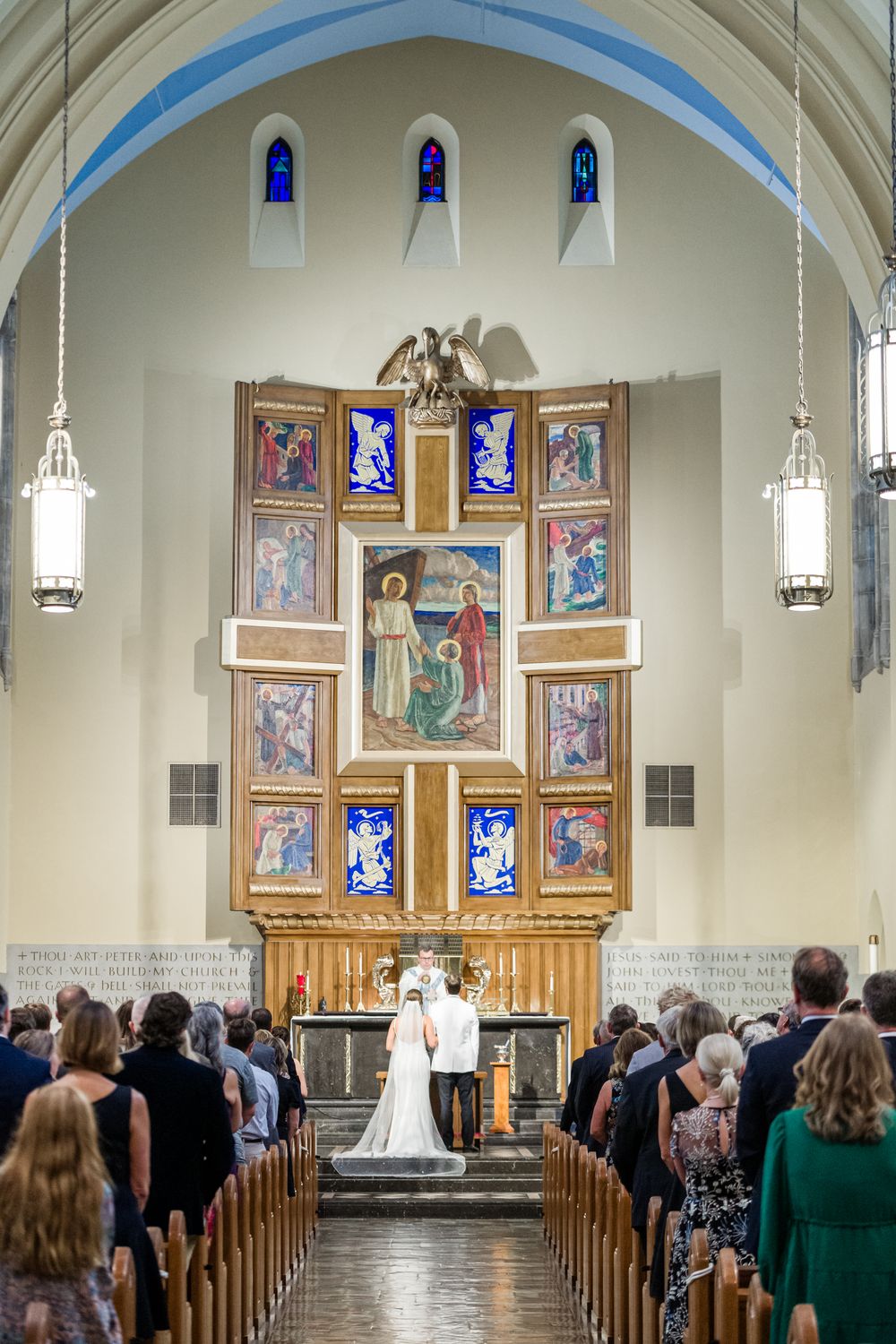 Couple exchanging vows at a church altar with ornate religious artwork and stained glass windows during wedding ceremony.
