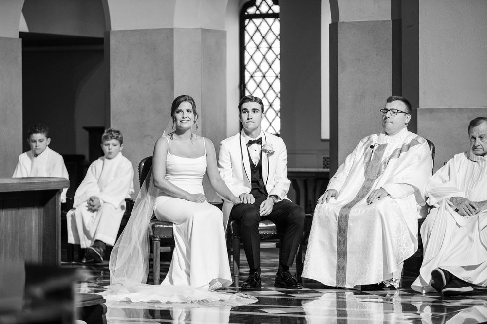 Black and white photo of a bride and groom seated at their wedding ceremony in a church with clergy nearby.