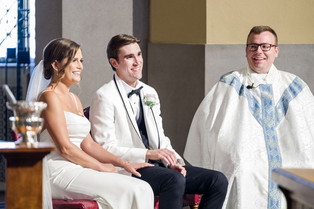 Bride and groom seated together smiling during wedding ceremony with officiant in white robes standing behind them.