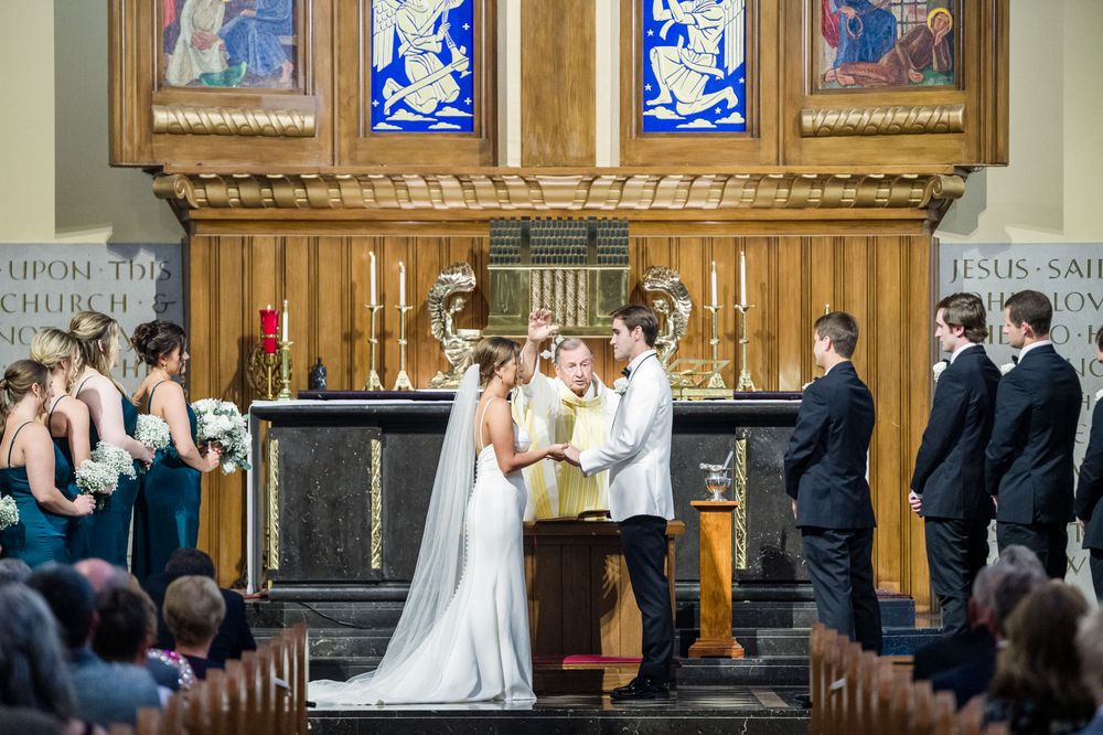 Bride and groom exchange vows at a church altar with stained glass windows, surrounded by wedding party.