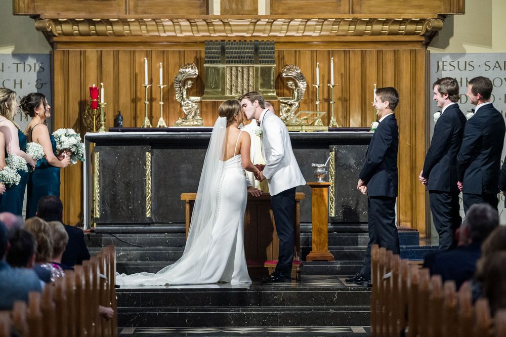 Bride and groom share a kiss at the altar during a church wedding ceremony with candles and wedding party.