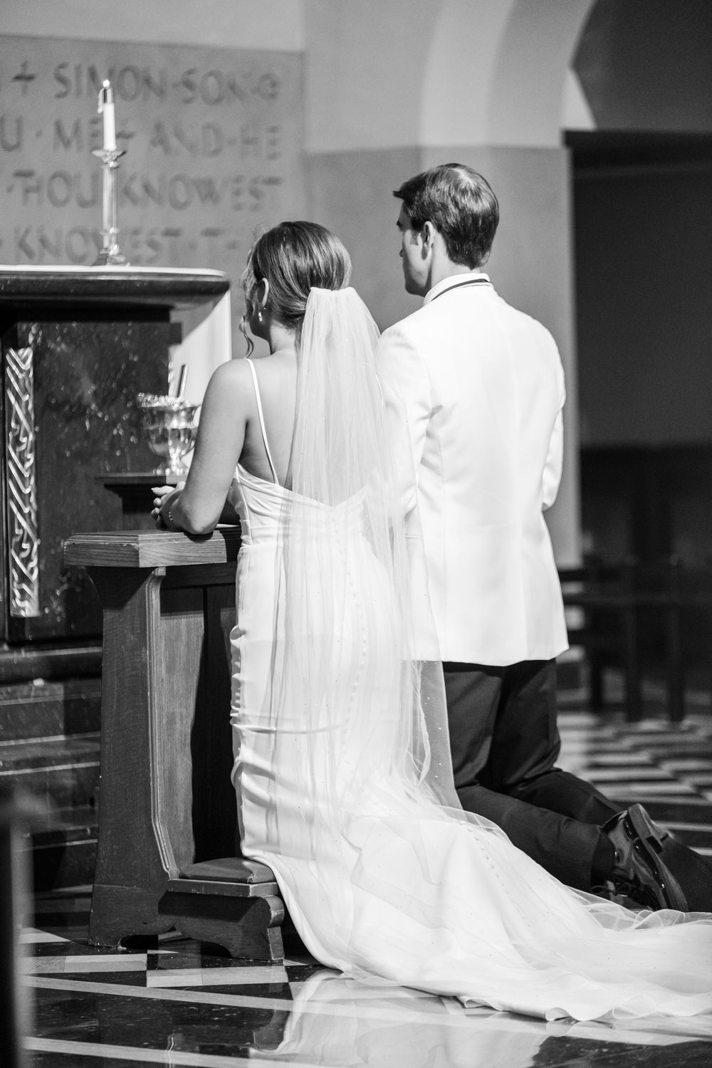Black and white photo of bride and groom kneeling at church altar during wedding ceremony.
