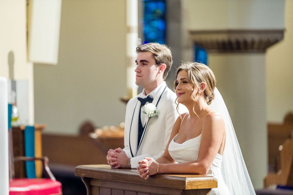 Bride and groom kneeling at church altar during wedding ceremony, bride in white gown, groom in white tuxedo.