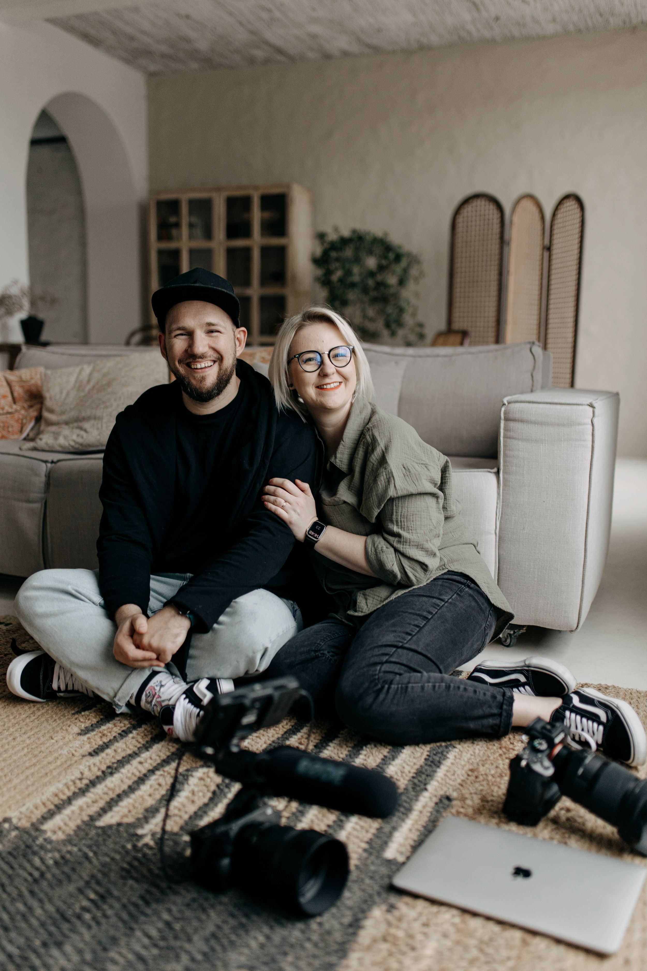 A smiling couple sits on a floor surrounded by cameras and a laptop in a cozy living room setting.