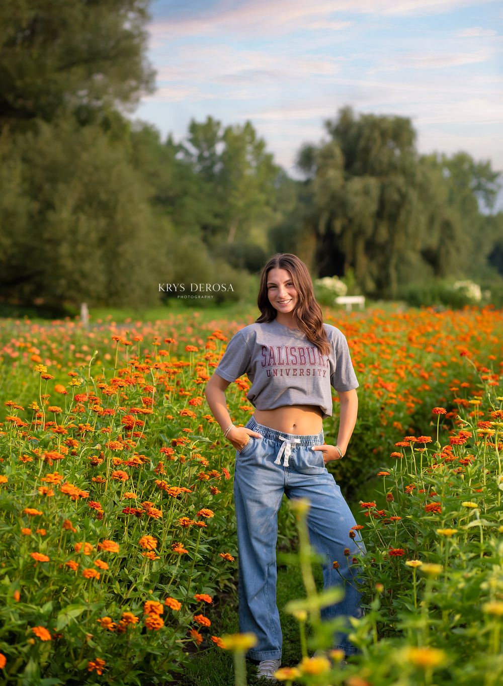 Young woman in gray Salisbury University crop top and jeans posing in vibrant orange flower field on sunny day.