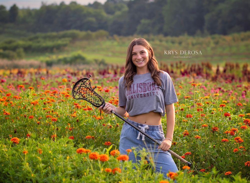 Teen girl holding lacrosse stick in vibrant wildflower field wearing Salisbury University shirt, senior portrait photography.