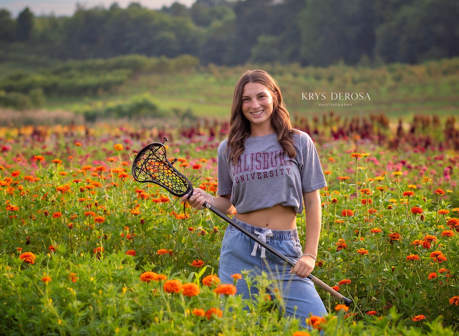 Teen girl holding lacrosse stick in vibrant wildflower field wearing Salisbury University shirt, senior portrait photography.