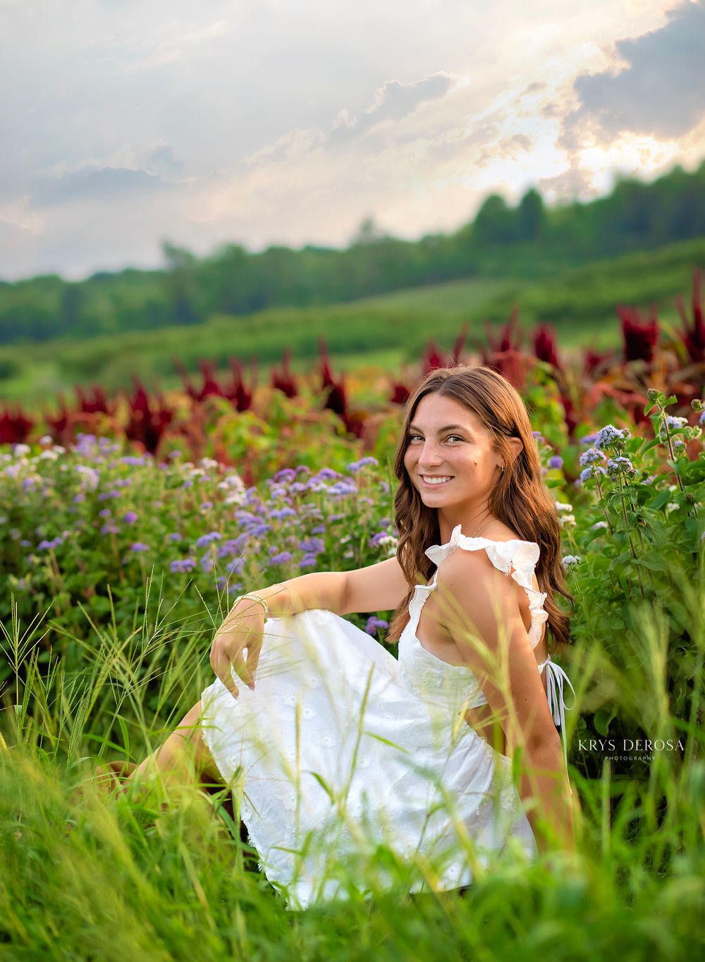 Young woman in white dress sitting among colorful wildflowers and red amaranth in a lush green field at sunset.