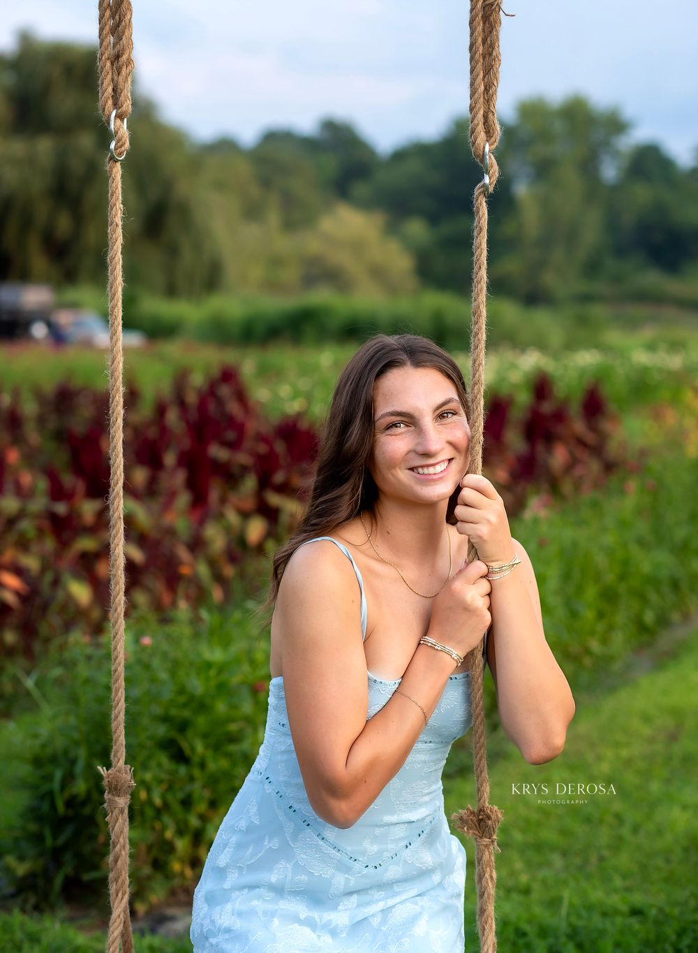 Smiling young woman in a light blue dress sitting on a rope swing surrounded by lush green and red gardens.