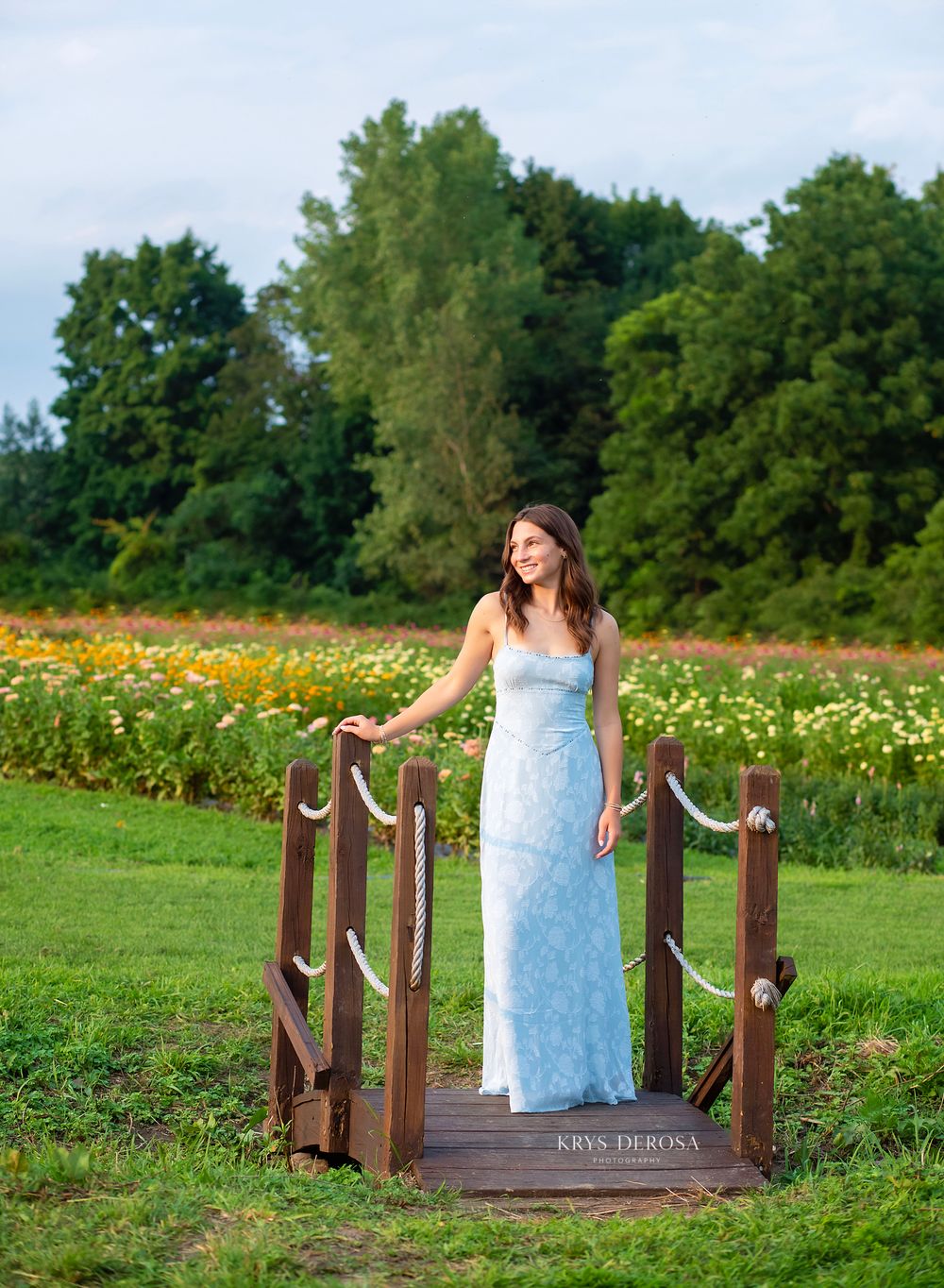 Young woman in light blue gown stands on wooden bridge surrounded by colorful flower fields and lush green trees.