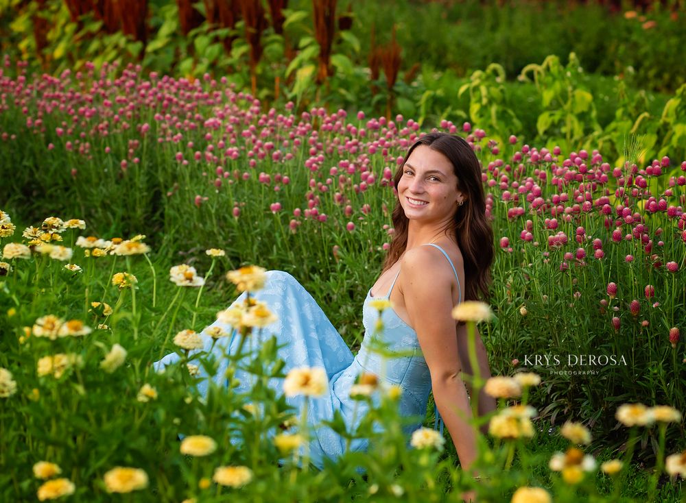 Smiling young woman in light blue dress sitting among colorful wildflowers in a lush garden setting.