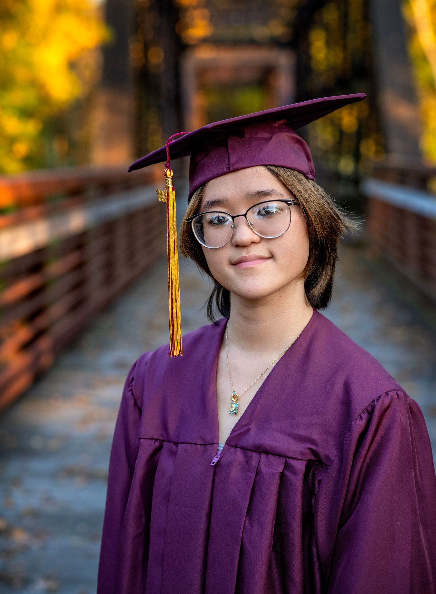 A young graduate smiles in maroon cap and gown on a bridge surrounded by autumn foliage.