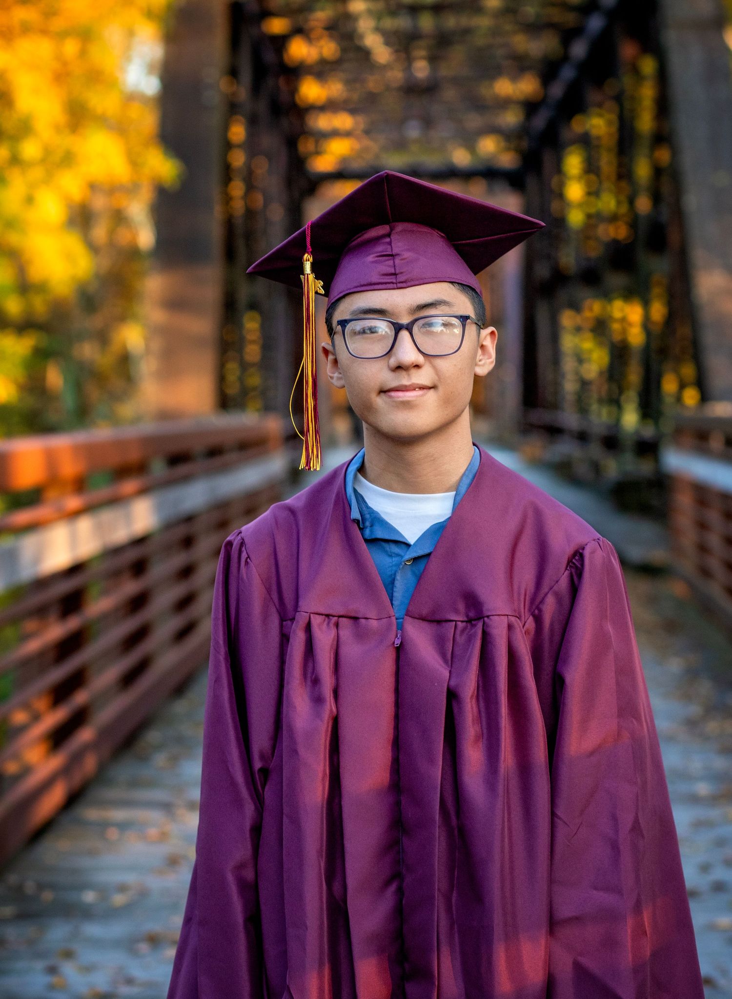 Young graduate in maroon cap and gown smiling on a wooden bridge surrounded by autumn foliage.