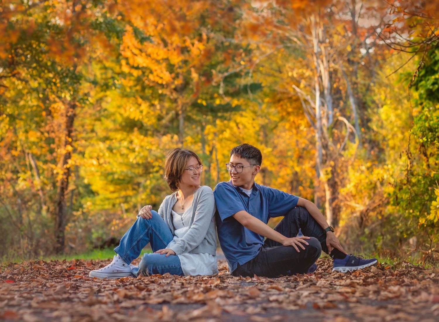 Couple sitting on fallen autumn leaves in a colorful forest with vibrant orange and yellow foliage.