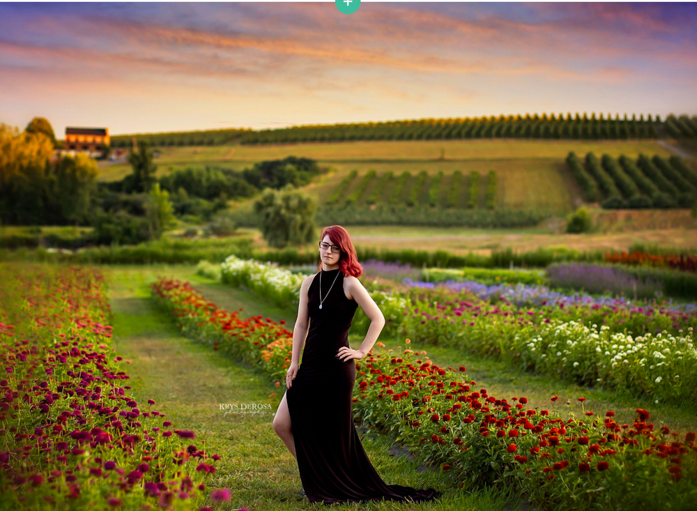 Woman in black gown poses in colorful flower garden with rolling green hills and dramatic sunset sky.
