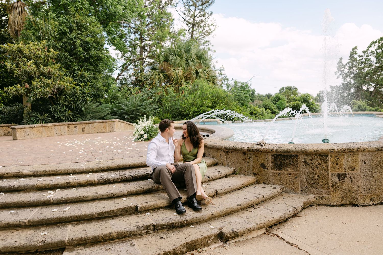 Couple sitting on stone steps by a garden fountain, surrounded by lush green trees, sharing a romantic moment outdoors.