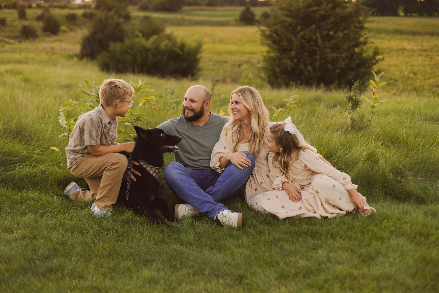 Family of four with a black dog sitting on lush green grass during golden hour in a scenic outdoor setting.