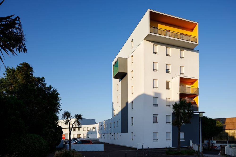 Immeuble résidentiel contemporain à plusieurs étages, doté de balcons aux couleurs vives jaune et orange, sous un ciel bleu éclatant.