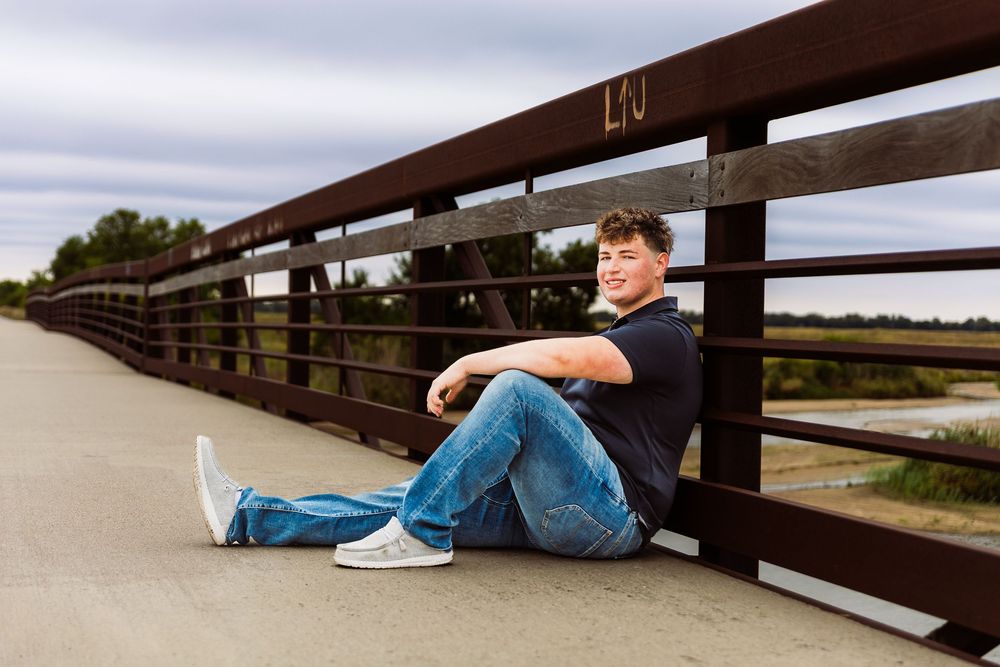 A smiling teenage boy sits against a wooden bridge railing on a paved path, wearing jeans and white sneakers.