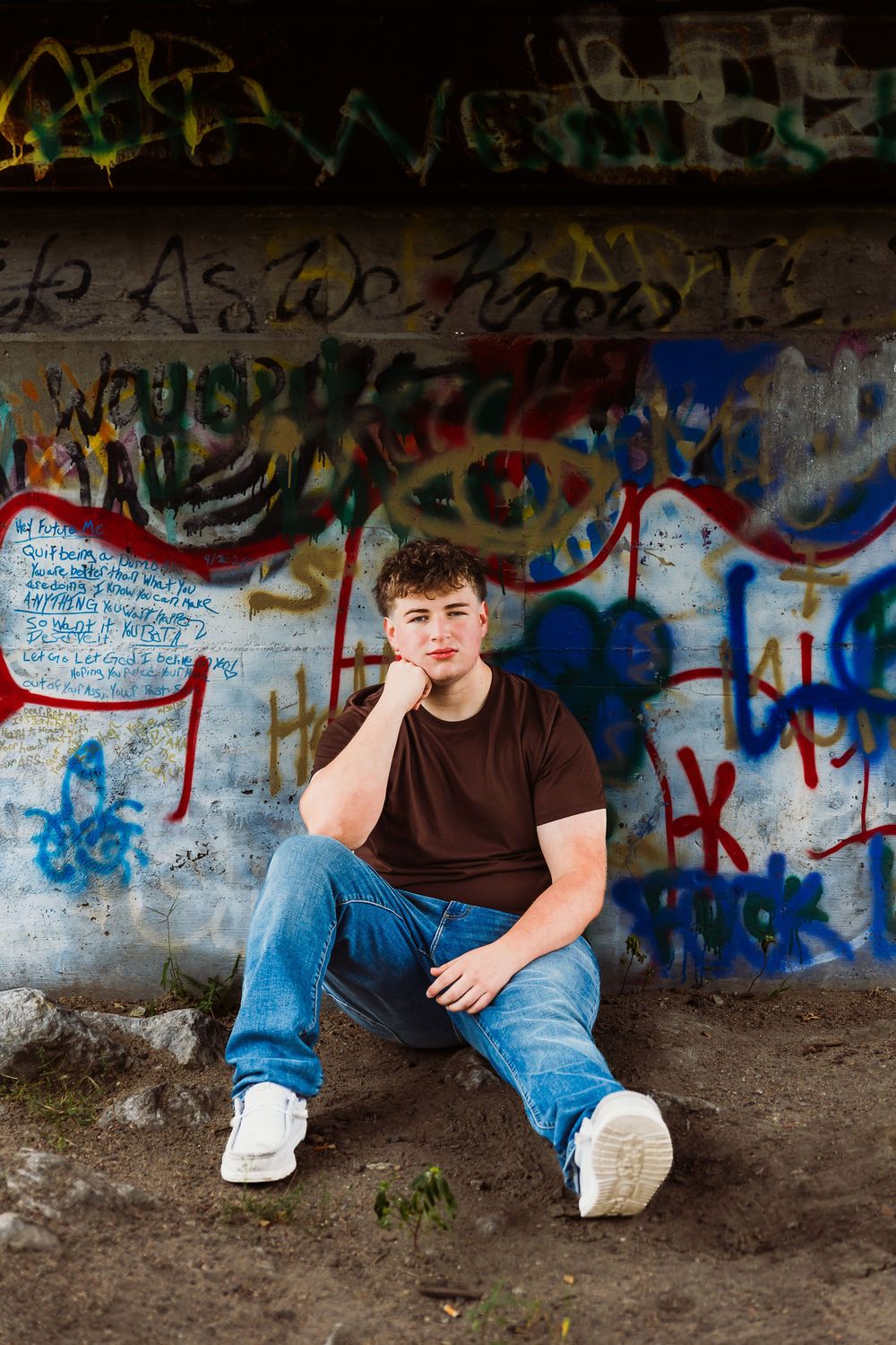 Young man in brown t-shirt and jeans sitting against a colorful graffiti wall outdoors.
