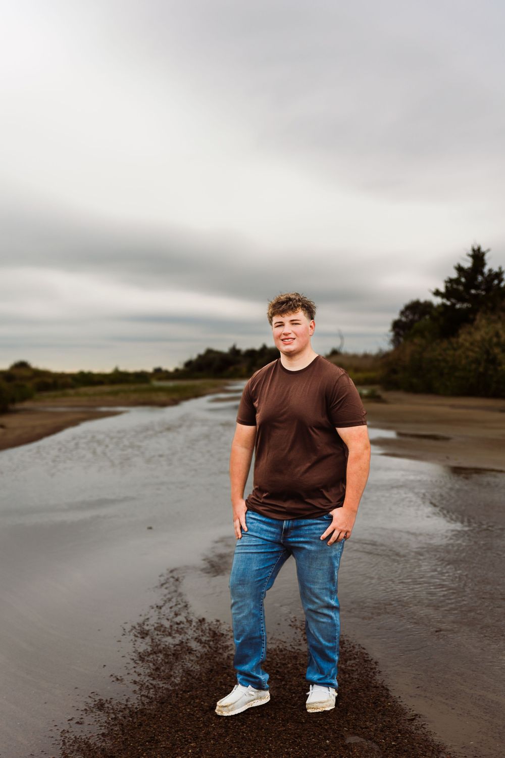 A teenage boy in a brown t-shirt and jeans stands confidently on a wet sandy beach under an overcast sky.