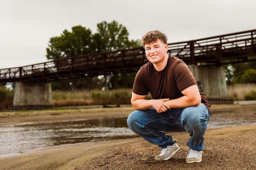 Young man crouching near a river with a bridge in the background, smiling outdoors.