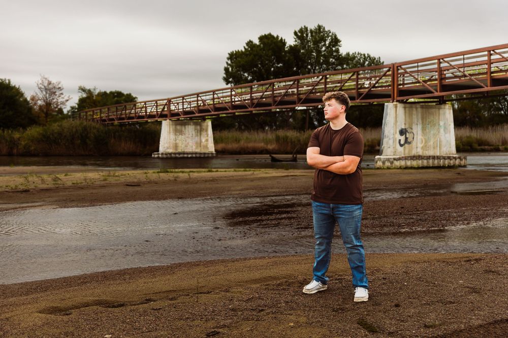 Man standing with arms crossed near a rustic bridge over a dry riverbed on a cloudy day.