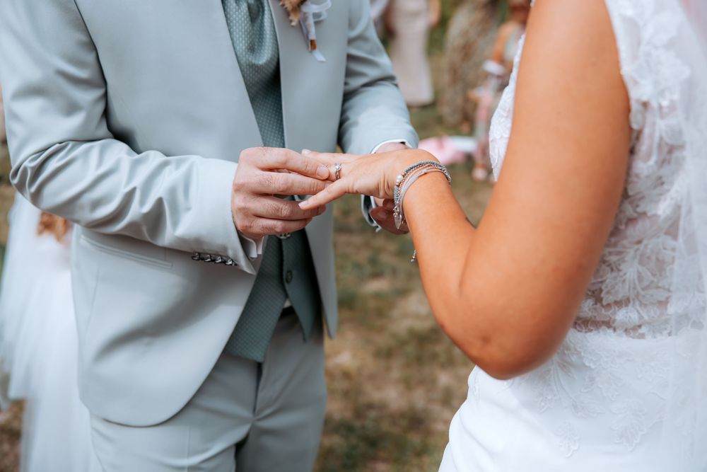 Groom in gray suit placing wedding ring on bride's finger during outdoor ceremony.