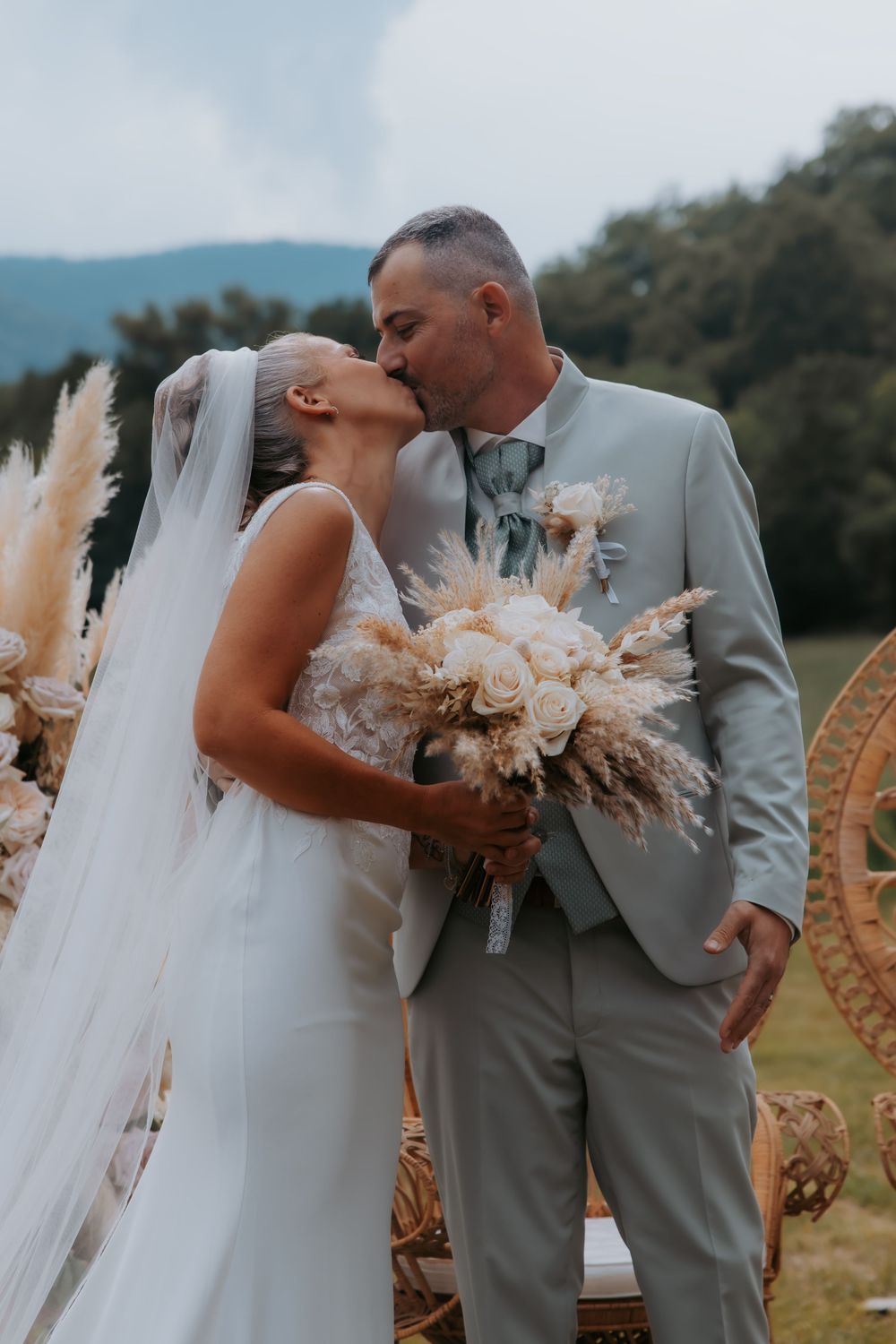 Bride and groom share a kiss at outdoor boho wedding ceremony, bride holding pampas grass bouquet in white gown.