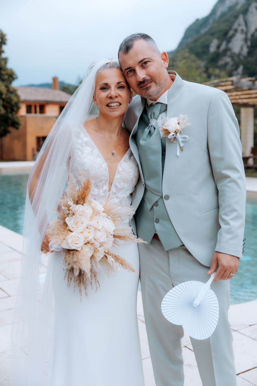 Bride and groom posing by a pool, she holds a boho bouquet in a lace gown, he wears a light blue suit.