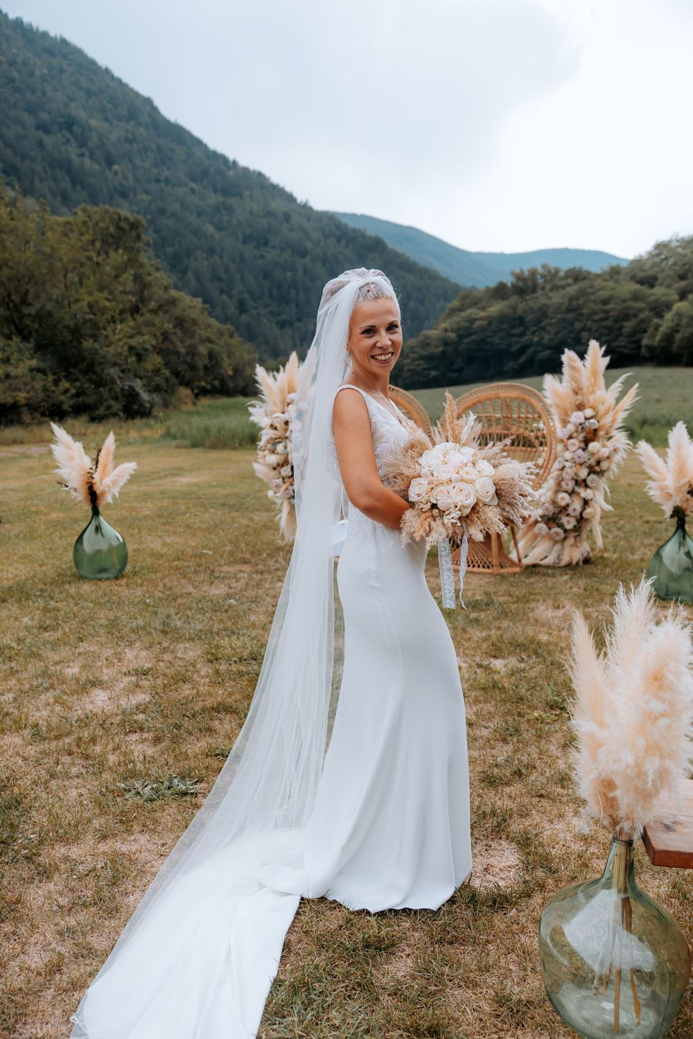 Smiling bride in white gown and veil holding bouquet outdoors with pampas grass decor and mountain backdrop.