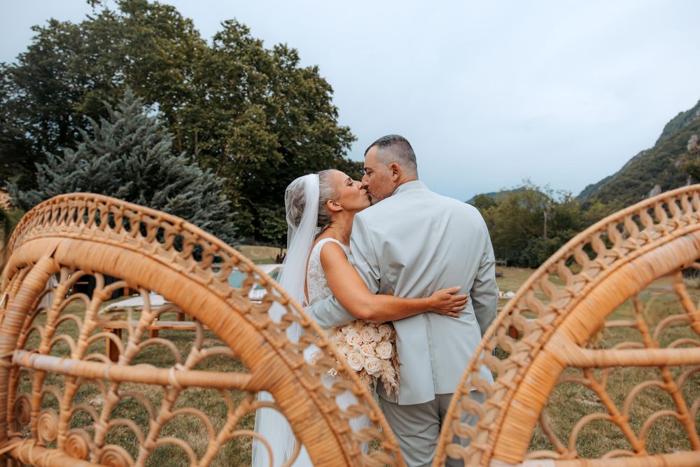 Bride and groom sharing a romantic kiss beside wicker chairs outdoors with lush green trees in the background.
