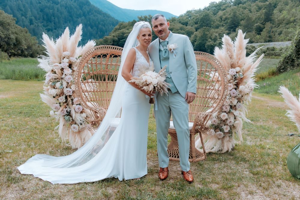 Bride and groom pose outdoors before a boho rattan peacock chair with pampas grass floral arch.
