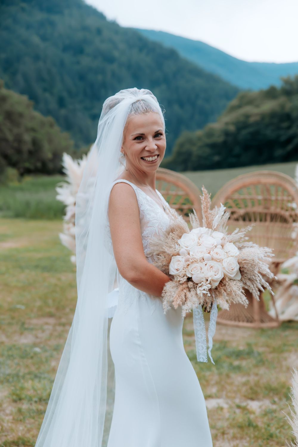 Smiling bride in white gown and veil holds boho bouquet with pampas grass, mountains and greenery in background.