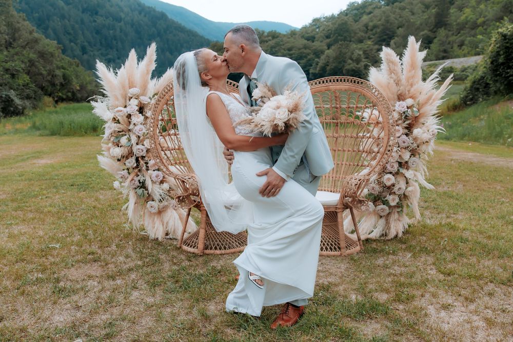 Bride and groom kissing in front of a boho rattan peacock chair adorned with pampas grass outdoors.