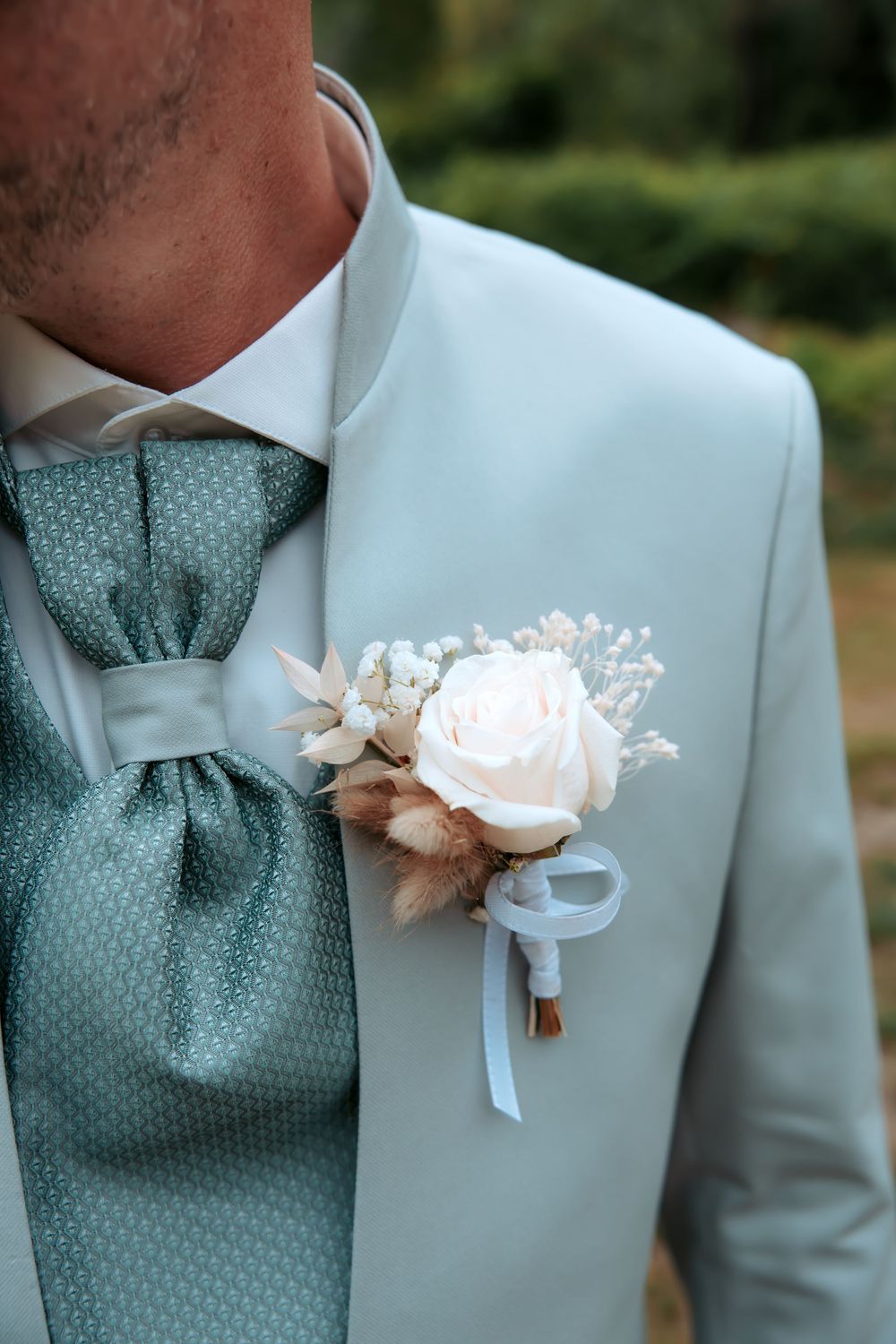 Groom wearing light blue suit with white rose boutonniere and sage green cravat tie at wedding ceremony.